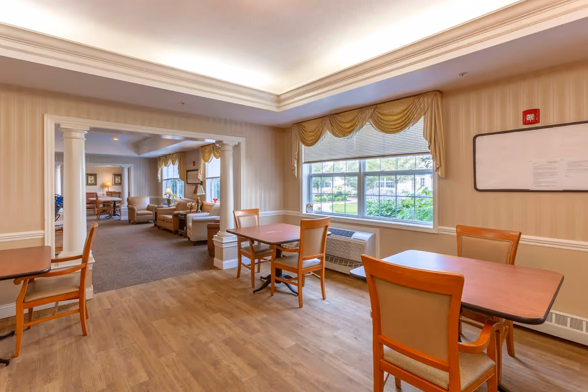 A well-lit interior room with wooden tables and chairs arranged near a large window with yellow curtains. The room has light-colored walls with white trim and a wooden floor. In the background, there is an adjoining lounge area with armchairs and a lamp, separated by white columns.