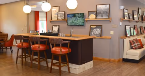 Interior view of a common area in Stonecroft Health Campus featuring a wooden bar counter with four high chairs with red cushions. The wall behind the bar has several framed pictures and a mounted flat-screen TV. There is a beige couch with a plaid pillow on the right side and additional seating visible in the background. The room has wooden flooring and warm lighting from hanging globe lights.