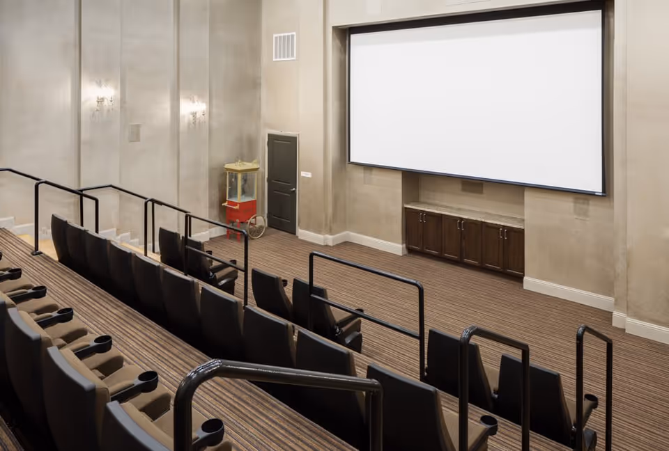An empty small theater room with tiered seating featuring black chairs with cup holders, a large blank projection screen on the front wall, beige walls, striped carpet, and a vintage popcorn machine in the corner.