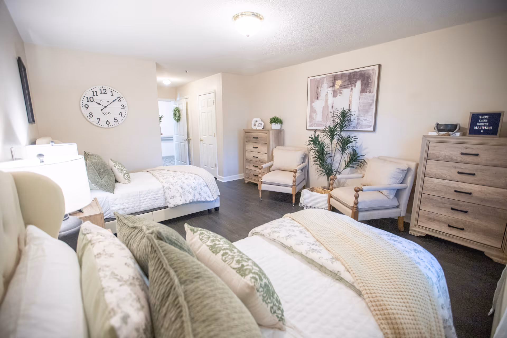 A bright and cozy bedroom with two beds, each made with white and patterned bedding and multiple pillows. The room features two wooden dressers, two cushioned armchairs, a large wall clock, a potted plant, and framed artwork on the walls. The floor is dark wood, and the walls are painted light beige.