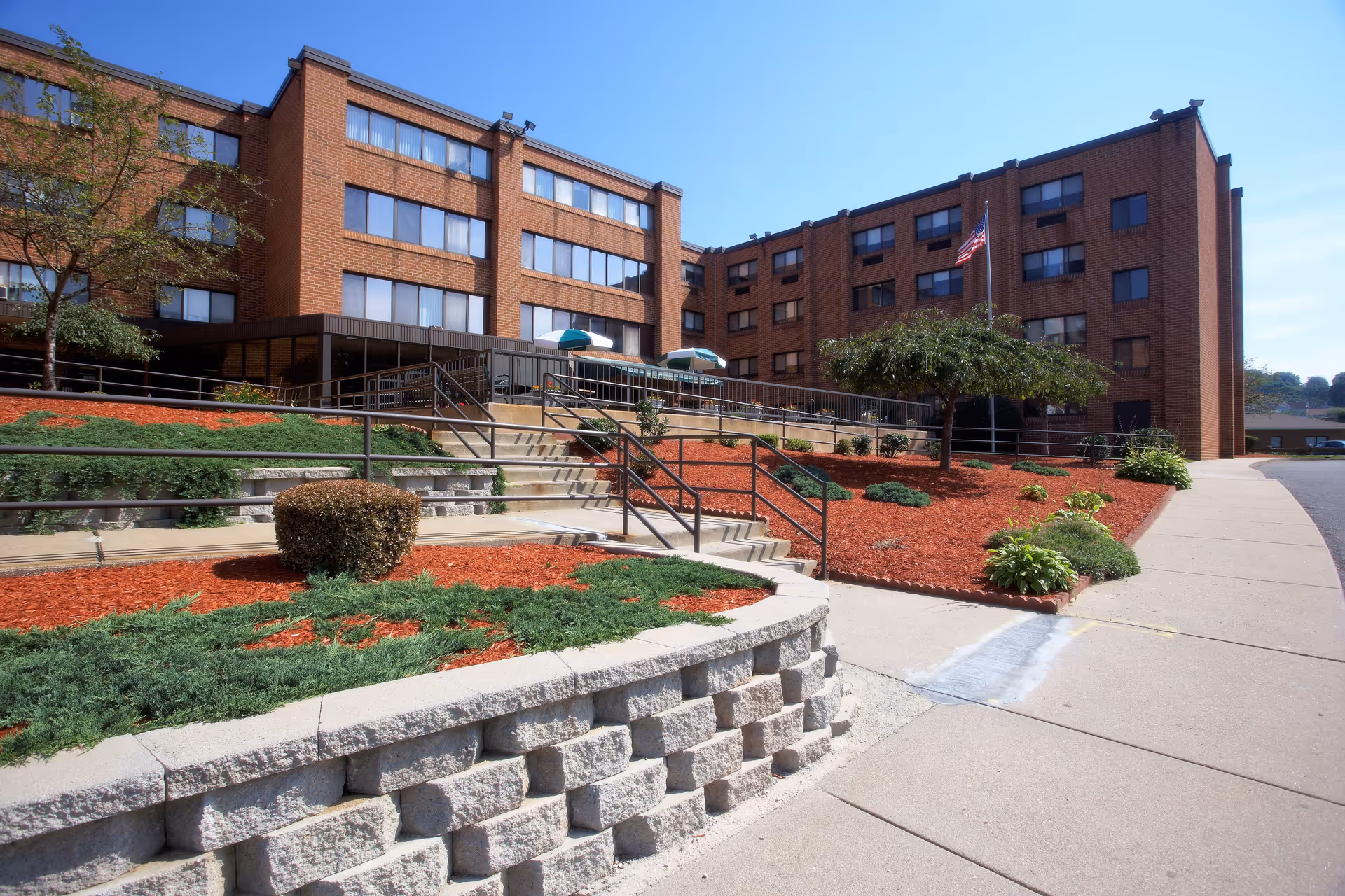 Exterior view of a multi-story brick building with several windows, a landscaped area with bushes and mulch, a concrete walkway, and stairs with metal railings leading to an entrance. An American flag is visible near the entrance under a clear blue sky.