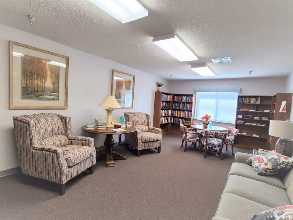 Cozy senior living lounge with armchairs, a sofa, bookshelves, and a round table.