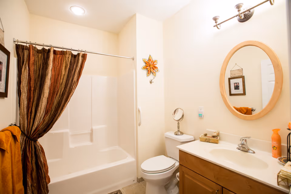 A clean bathroom with a white bathtub and shower combination, a brown and orange striped shower curtain, a white toilet, a wooden vanity with a white countertop and sink, an oval wooden-framed mirror above the sink, a wall-mounted light fixture, and orange towels and decor.