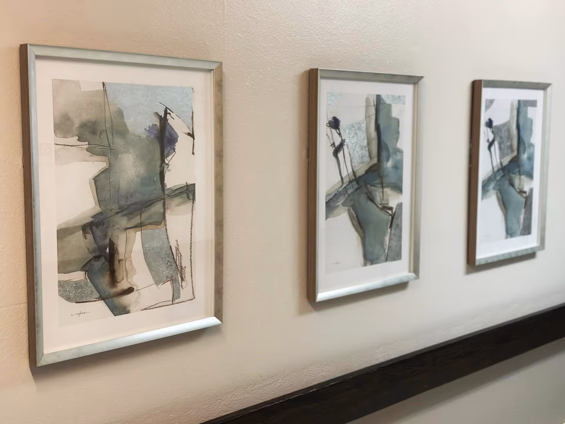 Three framed abstract watercolor paintings with shades of blue, gray, and beige hanging on a beige wall above a dark wooden chair rail.