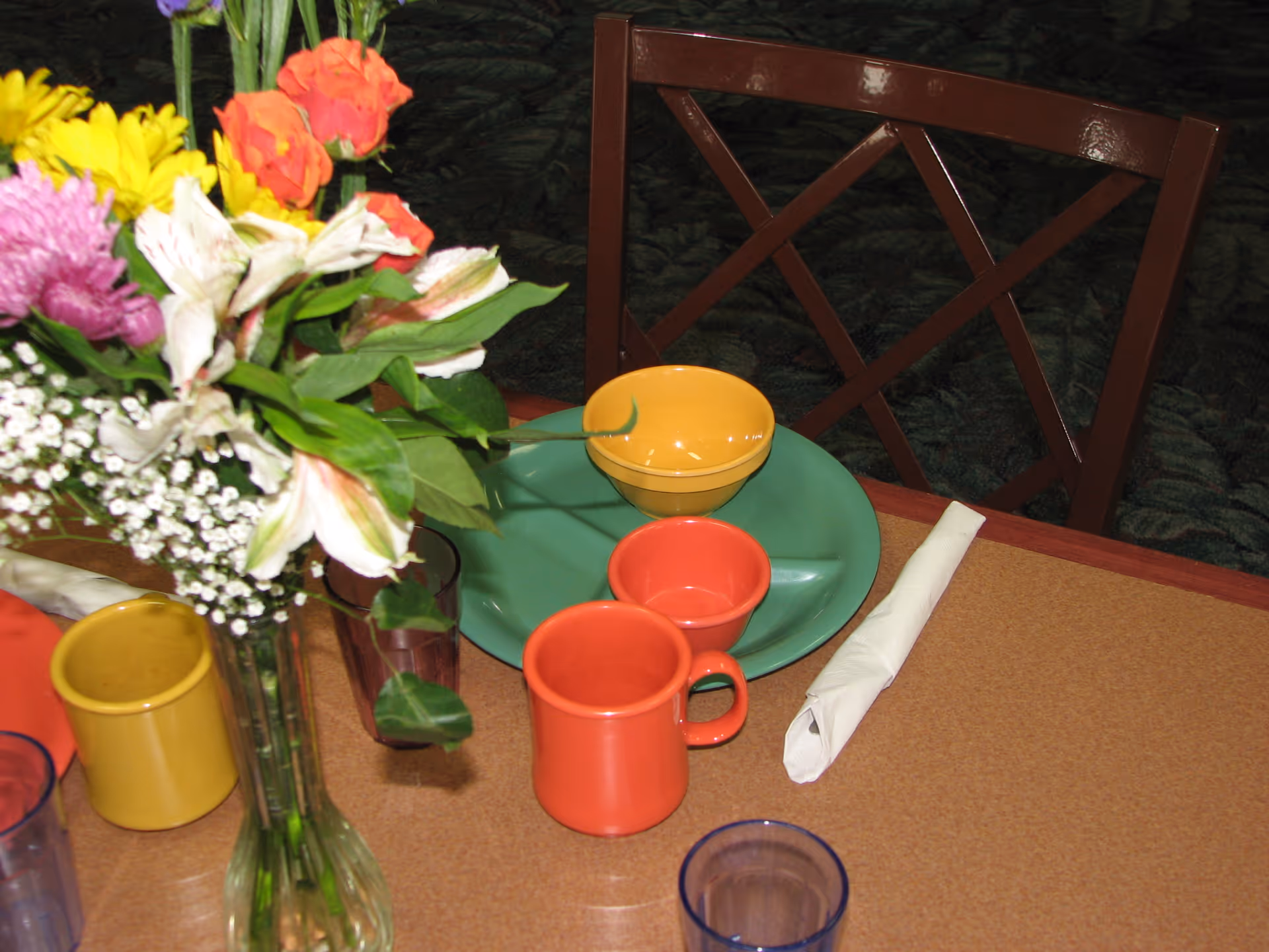 A dining table set with colorful dishes including a green plate, yellow bowl, orange cup, and orange mug. There is a rolled white napkin and a clear glass on the table. A vase with a bouquet of mixed flowers including yellow, orange, pink, and white blooms is also on the table. A wooden chair with a crisscross backrest is visible behind the table.