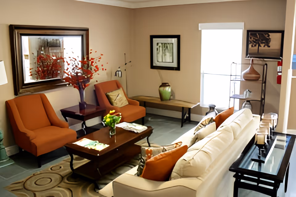 A cozy living room with a white sofa adorned with orange and patterned pillows, two orange armchairs, a wooden coffee table with a small flower vase, a side table with a lamp, a large mirror on the wall, a bench with a decorative vase, framed artwork, and a shelving unit with decorative items. The room has beige walls and a patterned rug on the floor.