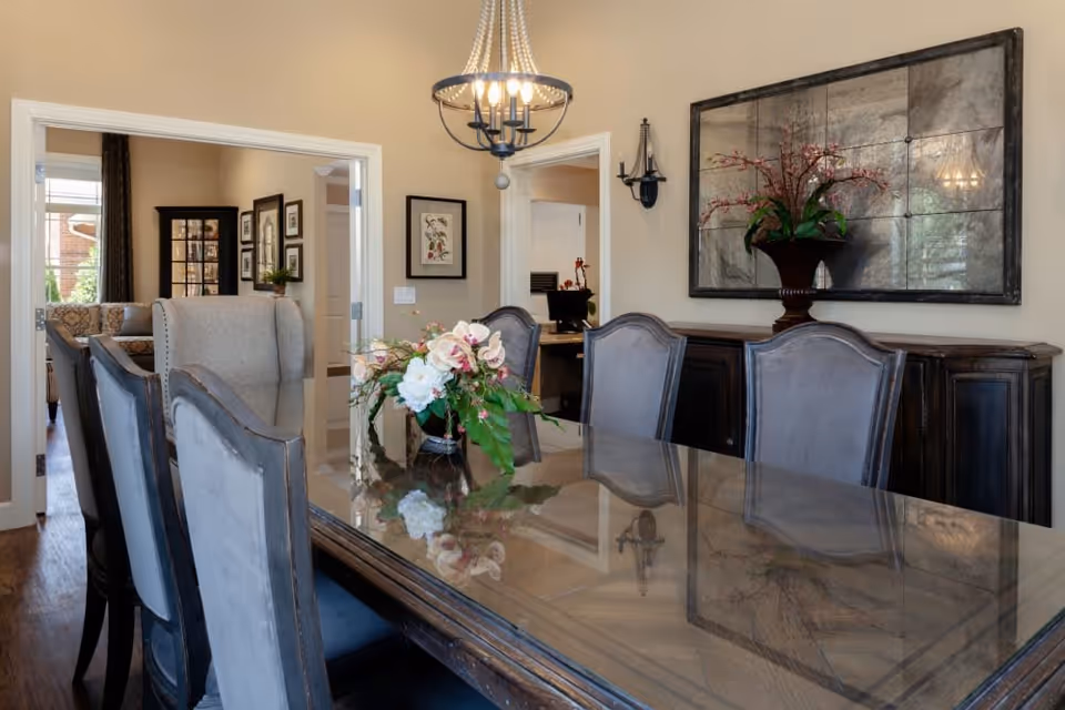 A dining room with a large wooden table and six upholstered chairs. A floral centerpiece is placed on the table. The room features a chandelier hanging from the ceiling, a large mirror on the wall, and a dark wooden sideboard beneath the mirror. In the background, there is an open doorway leading to another room with framed pictures on the wall.