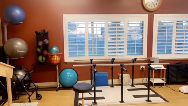 A therapy/exercise room with parallel bars, exercise balls, a walker, and a window with white shutters.