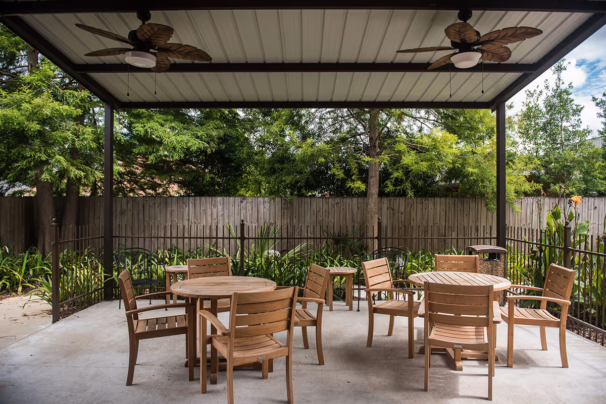 Outdoor covered patio area with two round wooden tables and several wooden chairs arranged around them. The patio has a metal roof with two ceiling fans featuring leaf-shaped blades. Surrounding the patio is a metal fence, and beyond that are green trees and plants with a wooden fence in the background.