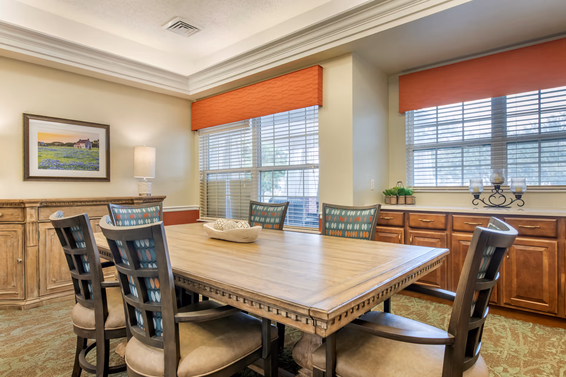 A dining room with a large wooden table surrounded by six cushioned chairs with patterned backs. The room has two large windows with orange valances, wooden cabinets along the walls, a table lamp, a framed landscape painting, and decorative items on the cabinets.
