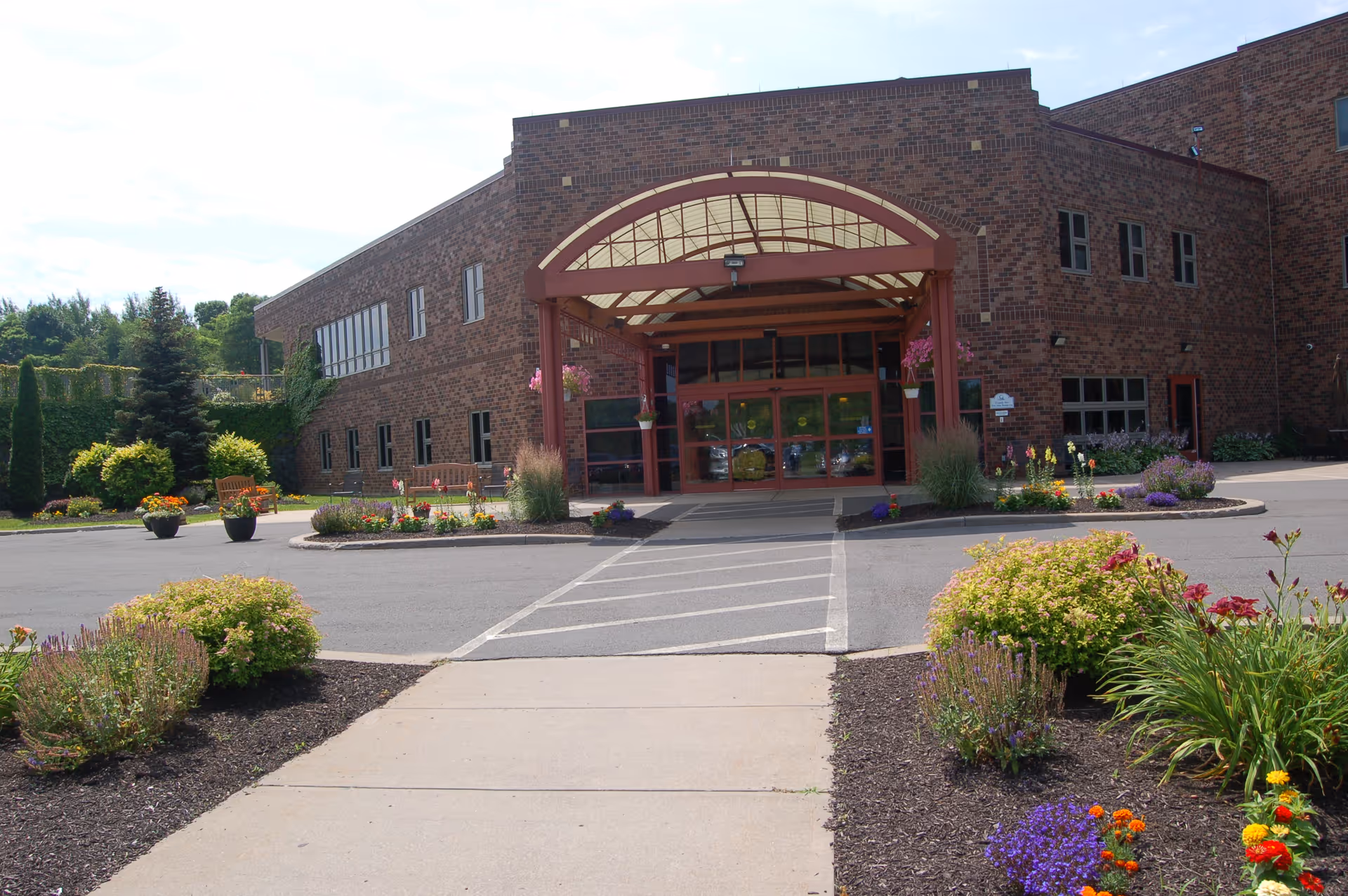 Front exterior view of a brick building with a covered entrance supported by red beams. There are landscaped flower beds with colorful flowers and shrubs on either side of the walkway leading to the entrance. The sky is partly cloudy.