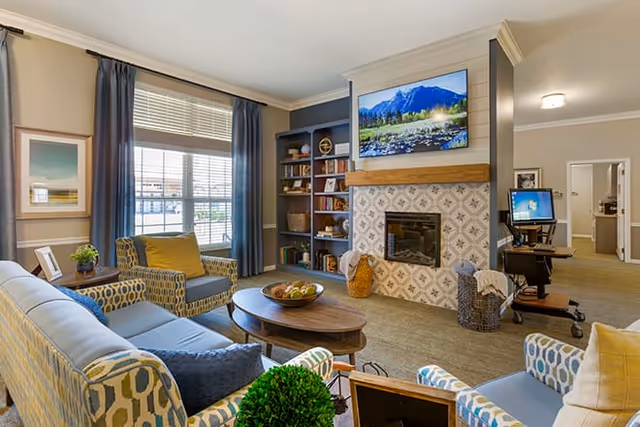 A cozy living room with patterned armchairs and a sofa arranged around a wooden coffee table. There is a fireplace with decorative tile and a flat-screen TV mounted above it. Built-in shelves next to the fireplace hold books and decorative items. A large window with blue curtains lets in natural light. In the background, a computer workstation is visible in an adjacent hallway.