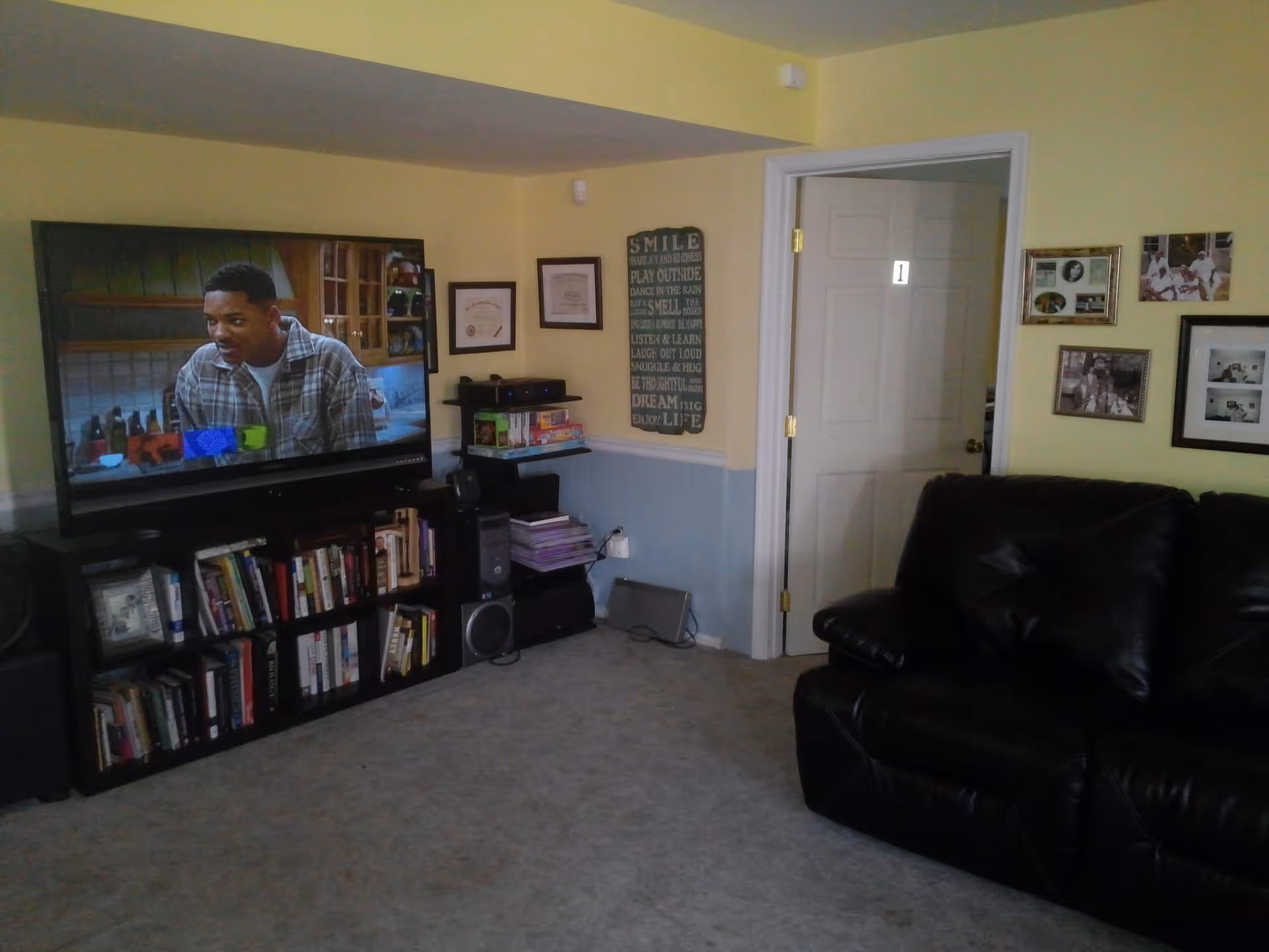 Living room with a large TV over a bookshelf, a black leather sofa, and framed photos on pale yellow walls.