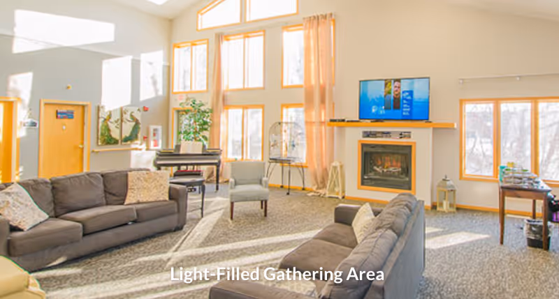 Bright communal living room with sofas, fireplace, TV, piano, and tall windows.