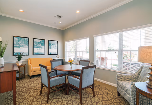 A bright and cozy common area with a round wooden table surrounded by four blue upholstered chairs. There is a yellow loveseat against the wall with three framed pictures above it. A patterned armchair and a wooden side table with a lamp are positioned near large windows letting in natural light. The room has beige walls and a patterned carpet.