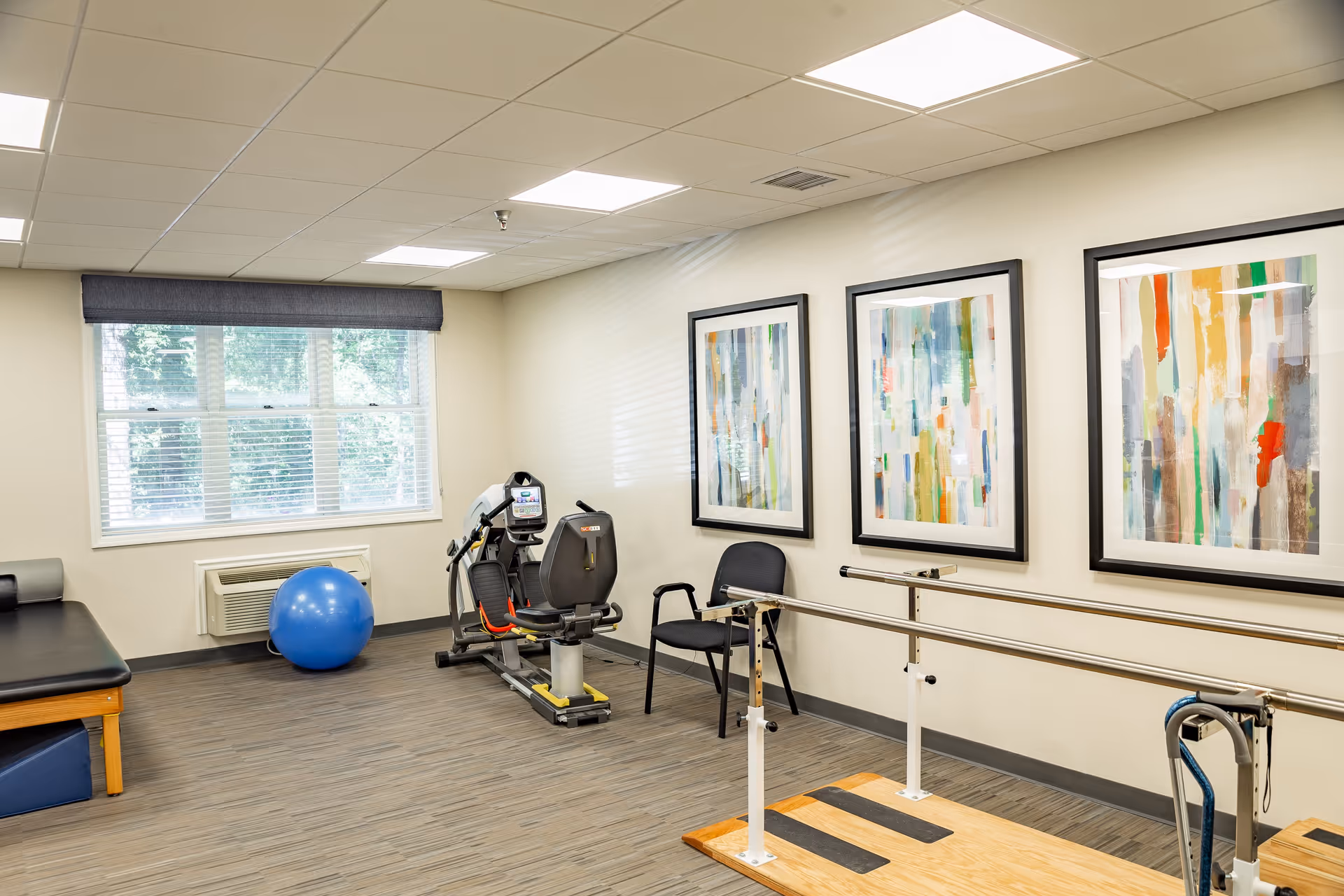 A bright therapy or exercise room with a large window, a blue exercise ball, a recumbent exercise bike, a black chair, parallel bars for walking support, and three colorful abstract paintings on the wall.
