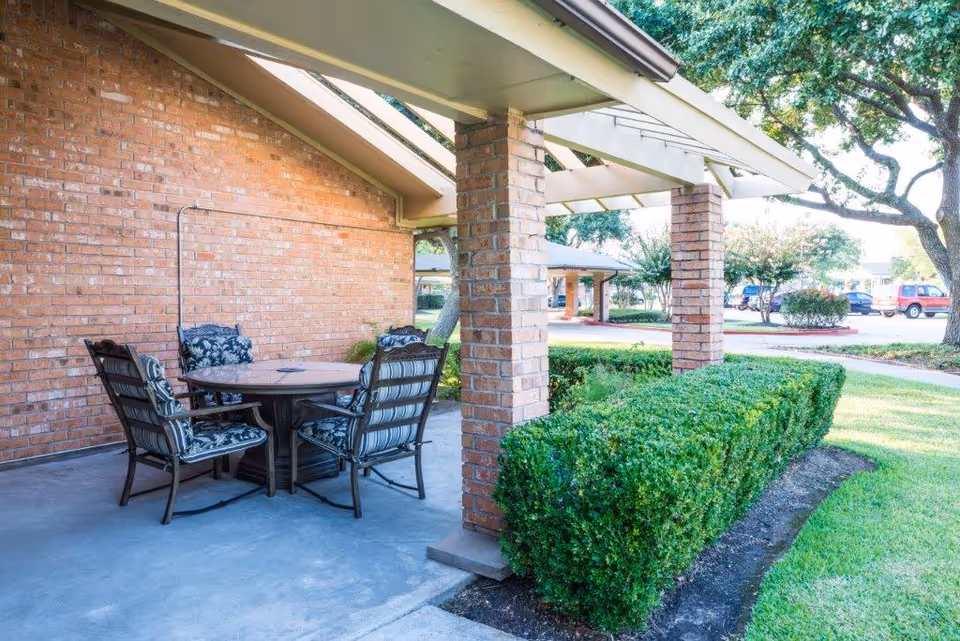 Outdoor covered patio area with a round table and four cushioned chairs, adjacent to a brick wall and surrounded by neatly trimmed bushes and trees in a landscaped garden.