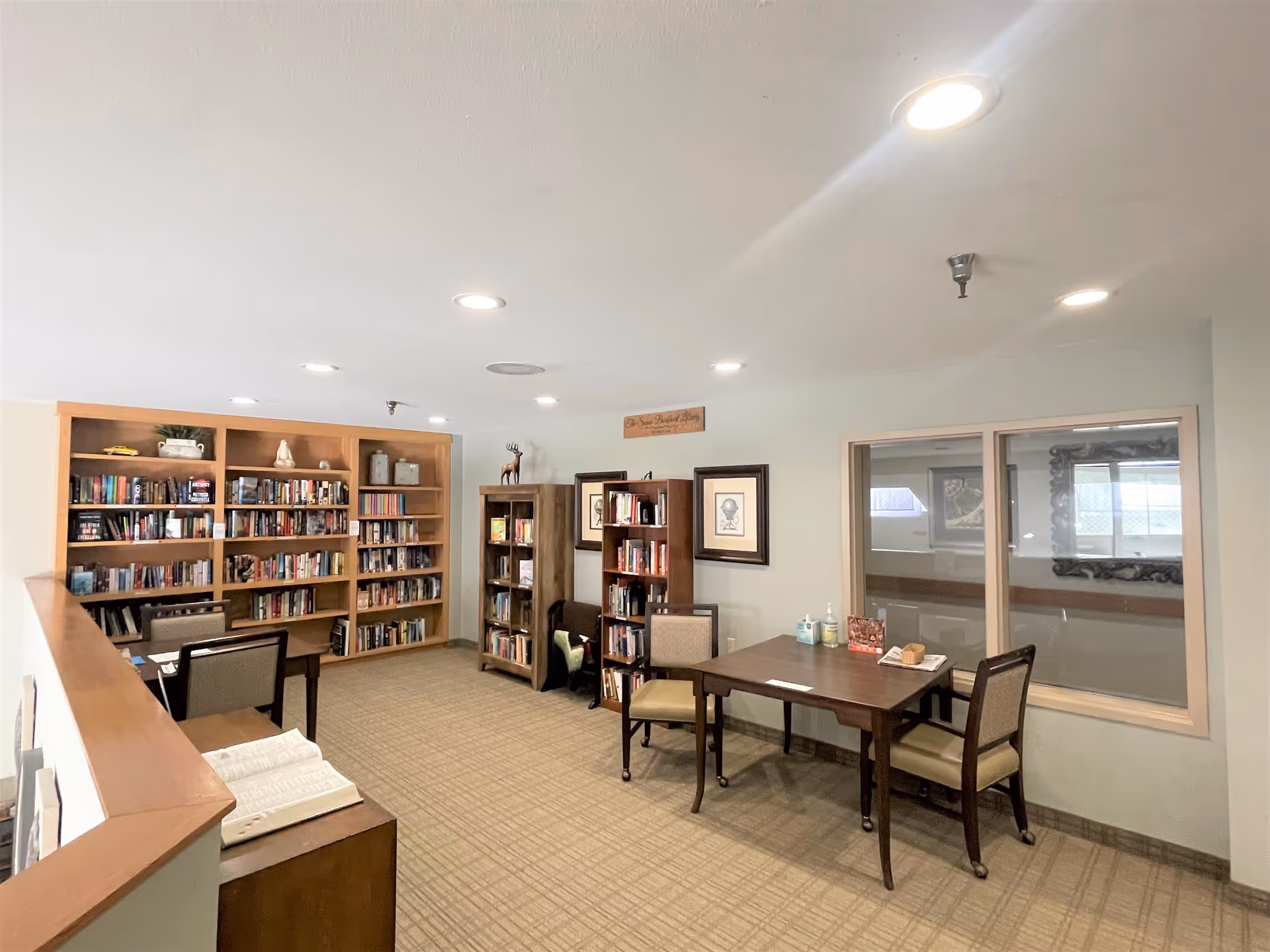 A quiet senior living facility library room with bookshelves filled with books, two tables with chairs, framed artwork on the walls, and a window looking into another room. The room is well-lit with recessed ceiling lights and has a carpeted floor.