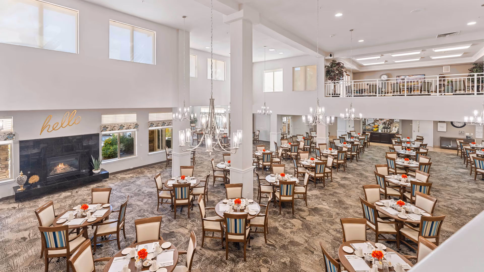 A spacious and well-lit dining room in a retirement living facility with multiple round tables set with white plates, cups, and red flower centerpieces. The room features high ceilings, large windows, chandeliers, a fireplace with the word 'hello' above it, and a second-floor balcony overlooking the dining area.