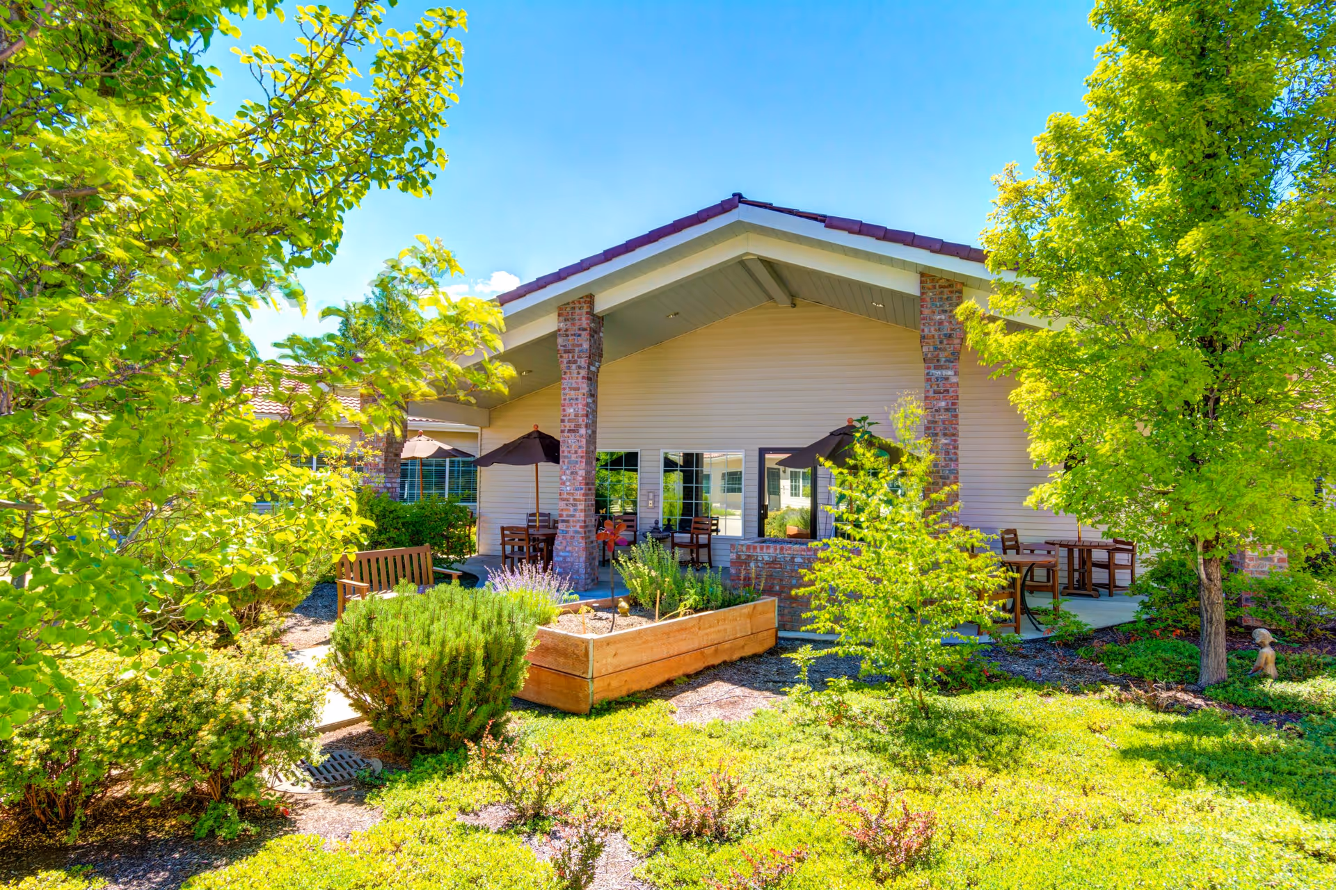 Outdoor patio area of a senior living facility with wooden benches, tables with umbrellas, surrounded by lush green trees and shrubs under a clear blue sky.