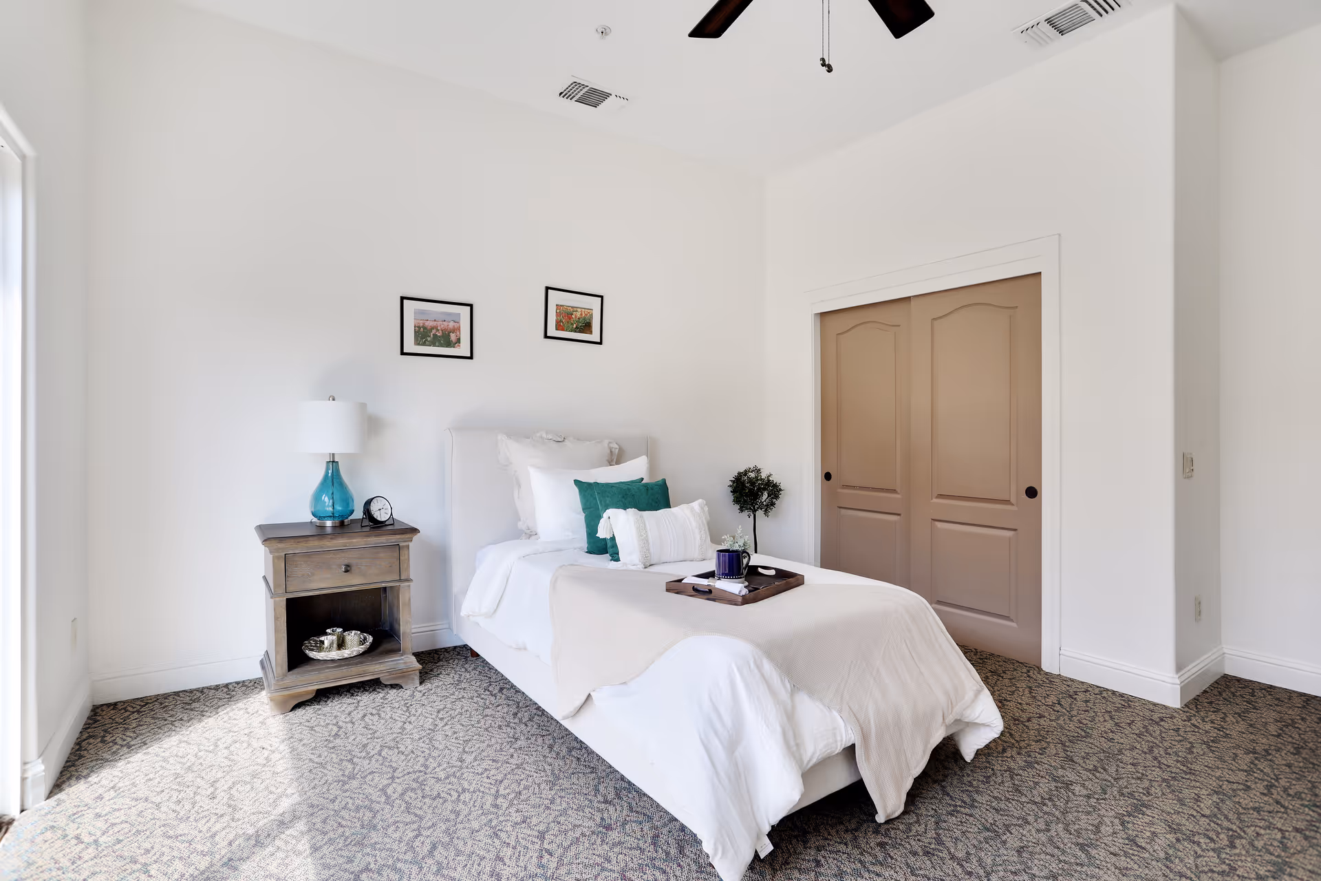 Bright single bedroom with a neatly made bed, bedside table with a lamp, and closed double closet doors.