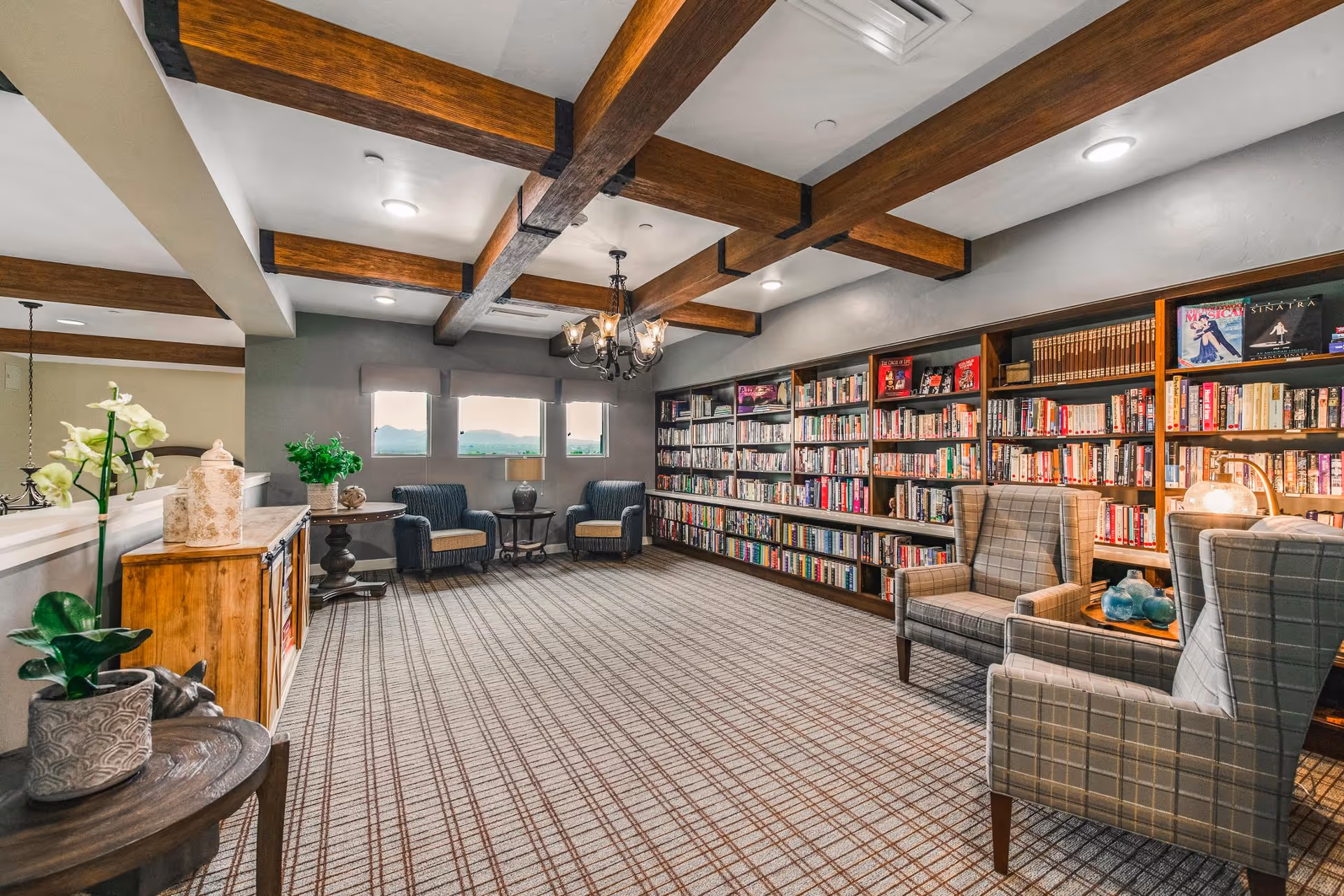 A cozy library room with wooden ceiling beams and a patterned carpet. The room features a large bookshelf filled with books along one wall, several comfortable armchairs arranged for reading, and small tables with decorative items and plants. Three windows allow natural light to enter the space.