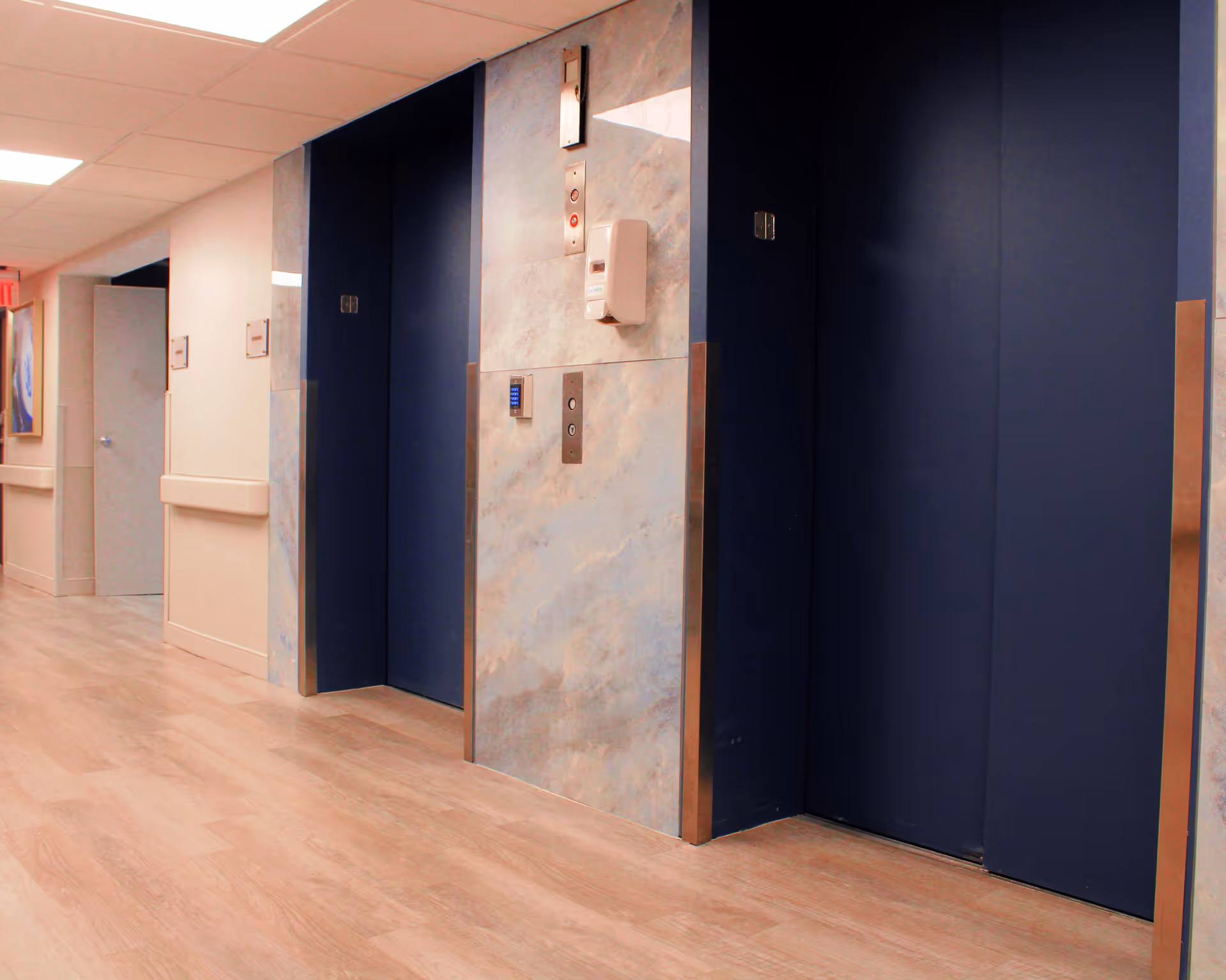Interior hallway of a facility with two closed blue elevator doors set in a wall with a marble-like finish. The floor is light wood, and the ceiling has recessed lighting. There are handrails along the walls and a partially open door at the end of the hallway.