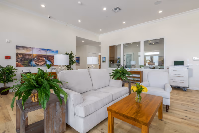 A bright and spacious living room area in a senior care facility featuring a light gray sofa and armchair, a wooden coffee table with a vase of yellow flowers, several green potted plants, two table lamps with white shades, and a multi-panel wall art depicting a scenic river bend. The room has light wood flooring, white walls, recessed ceiling lights, and large windows looking into an adjacent room.
