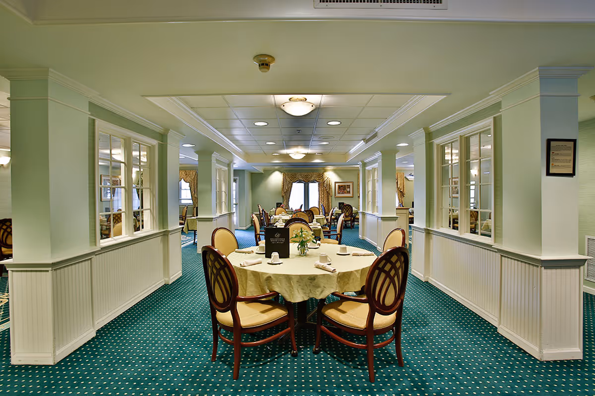 A bright dining room with round tables set for meals and upholstered chairs in a senior living facility.