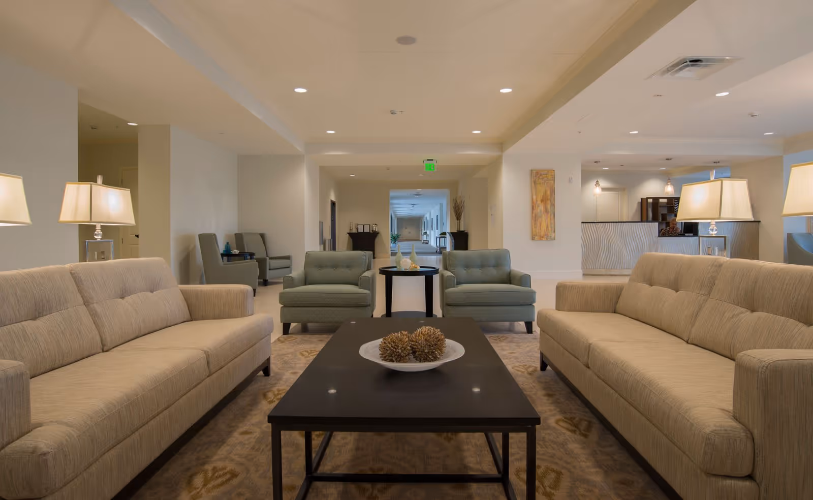 A spacious and well-lit living room area in The Blake at Flowood facility featuring two beige sofas facing each other with a dark wooden coffee table in between. The coffee table holds a decorative bowl with pine cones. Behind the sofas are two green armchairs with a small round table between them. The room has soft lighting from table lamps and recessed ceiling lights, with a hallway and reception area visible in the background.