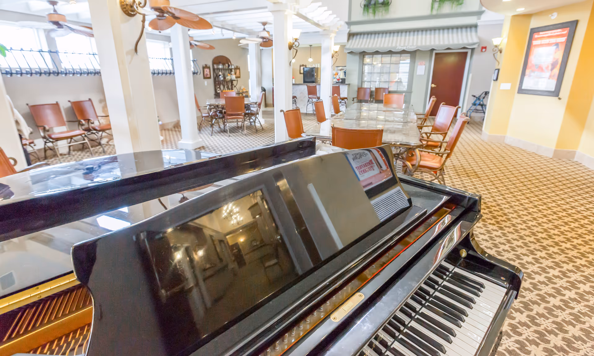 Interior view of a senior living facility common area featuring a black grand piano in the foreground, several tables with chairs arranged around the room, ceiling fans, and warm lighting. The room has patterned carpet and a cozy, inviting atmosphere.