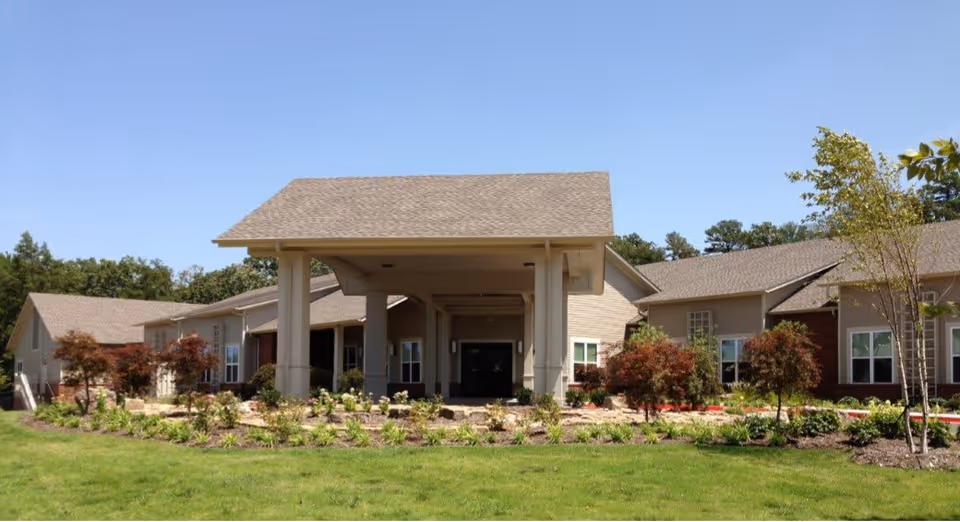Exterior view of Pinnacle Place Memory Care facility showing a single-story building with a large covered entrance, surrounded by landscaped greenery and small trees under a clear blue sky.