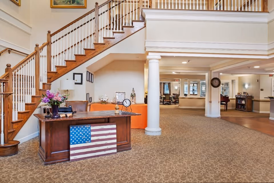 Interior view of a senior living facility lobby with a wooden reception desk adorned with a vase of flowers and a decorative American flag. Behind the desk is a staircase with wooden steps and white railings leading to an upper floor. The area has beige patterned carpet, white walls, and a large white column. In the background, there are seating areas and a clock mounted on a pillar.