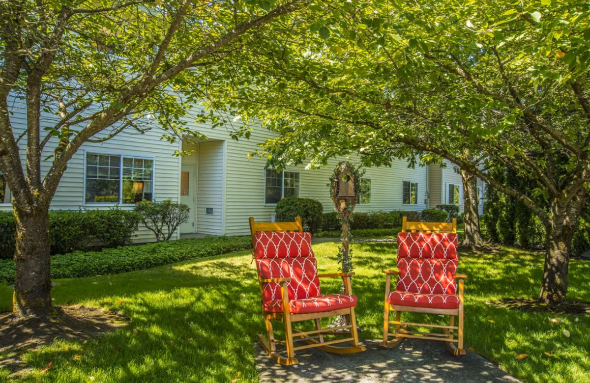Two wooden rocking chairs with red patterned cushions placed on a shaded concrete patio under leafy green trees in a grassy garden area outside a white building with multiple windows and bushes.