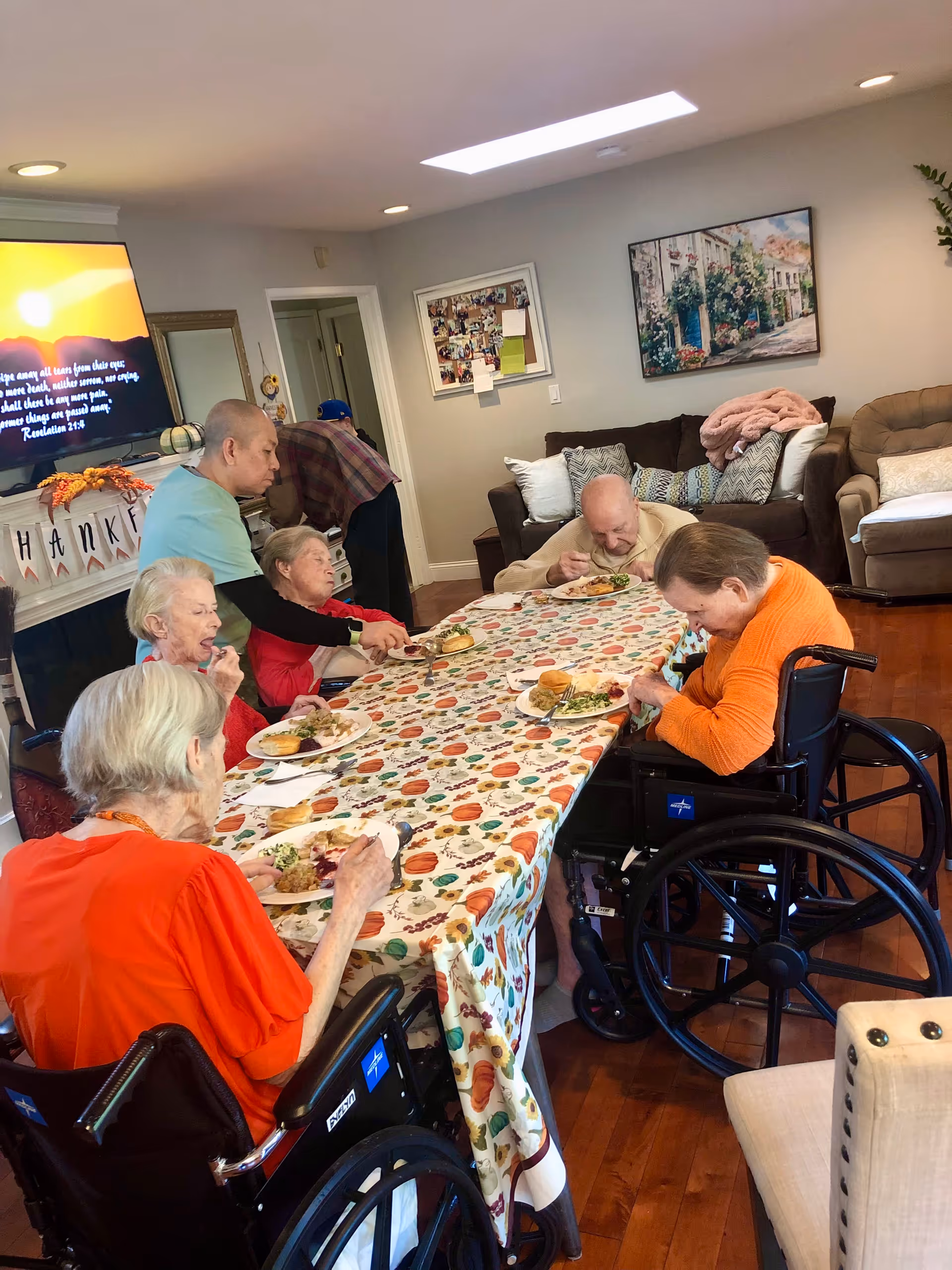 Elderly residents sit around a long festive table in a senior living dining area eating a meal.