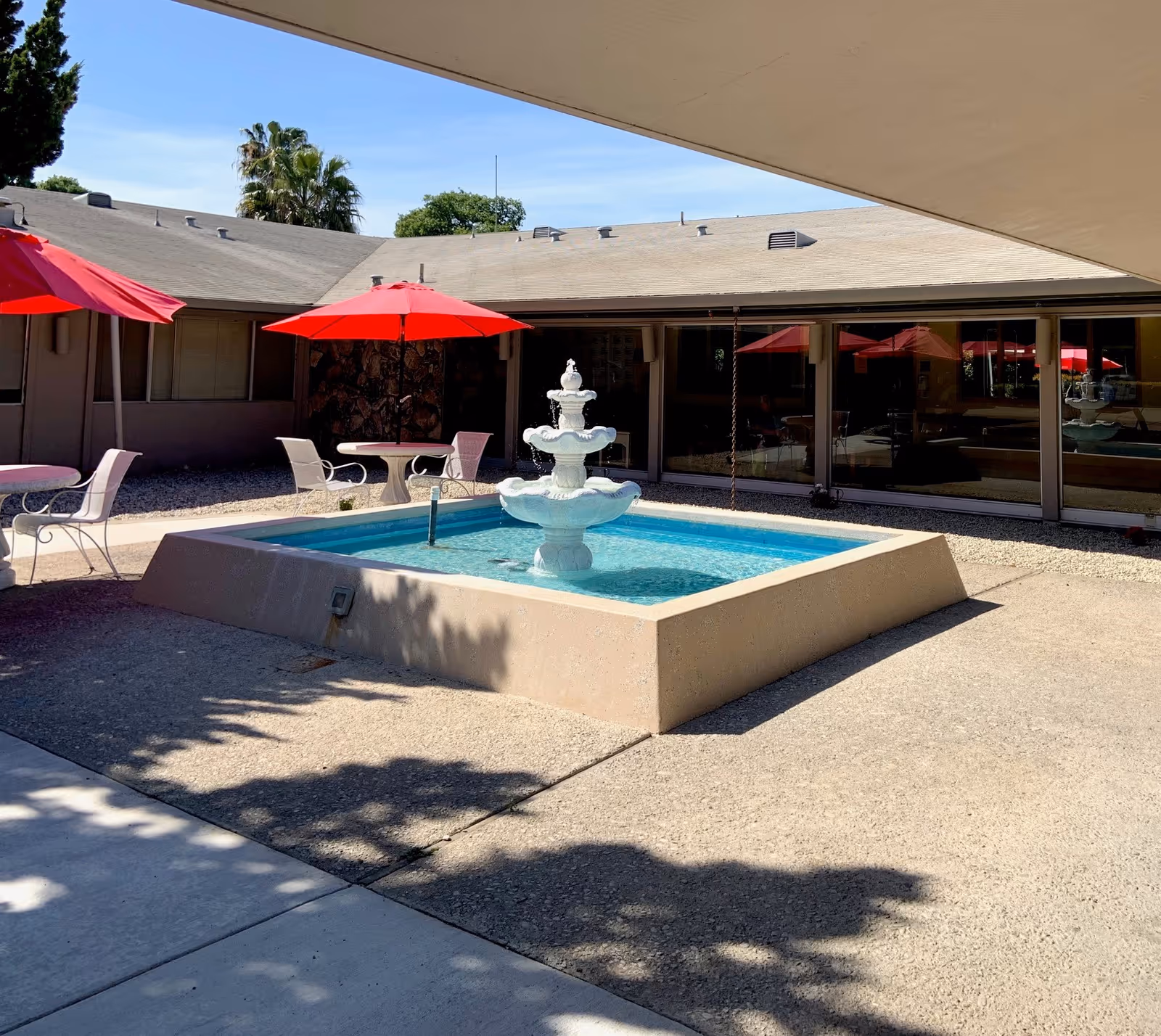 Outdoor courtyard area with a three-tiered white fountain in the center of a square pool. Surrounding the fountain are tables with red umbrellas and white chairs. The courtyard is enclosed by a single-story building with large windows reflecting the umbrellas and fountain. Trees and a clear blue sky are visible in the background.