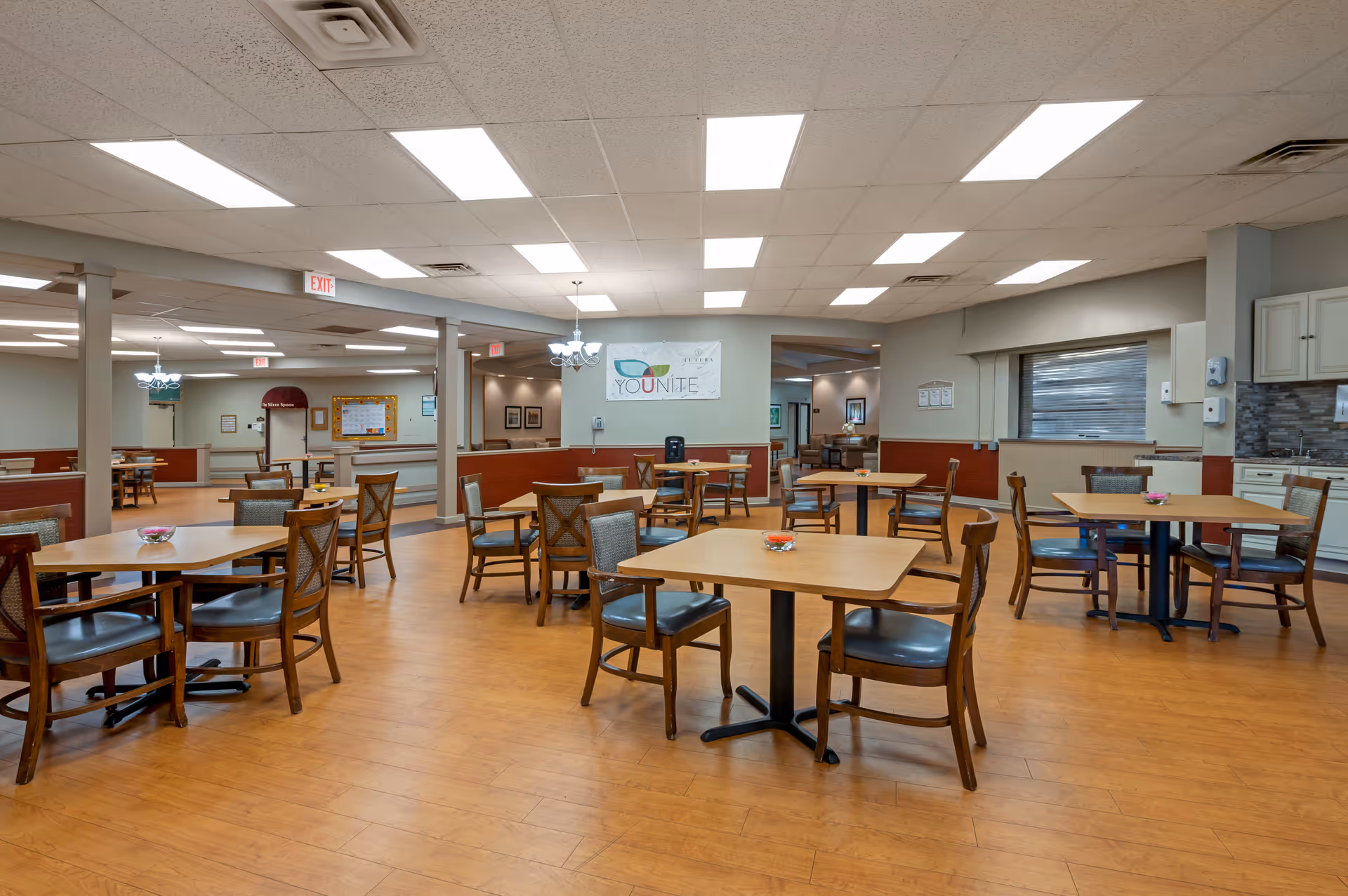Spacious dining room with multiple wooden tables and chairs in a senior care facility.