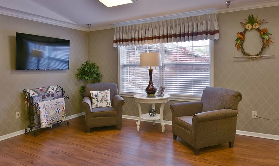 A cozy sitting area in Henley Place featuring two brown upholstered armchairs with floral pillows, a white round side table with a lamp and a framed photo, a wall-mounted flat-screen TV, a quilt rack with colorful quilts, a potted plant, and a window with blinds and a valance. A decorative wreath hangs on the wall.