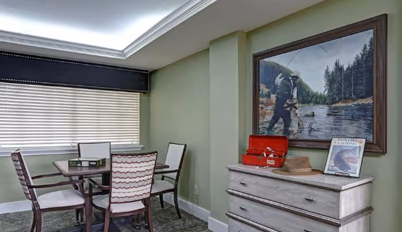 A small dining area with a square wooden table and four chairs with patterned upholstery. Next to the table is a light-colored chest of drawers with a red tackle box, a brown hat, and a framed magazine titled 'Fishing' on top. Above the chest hangs a large framed painting of a person fly fishing in a river surrounded by trees.