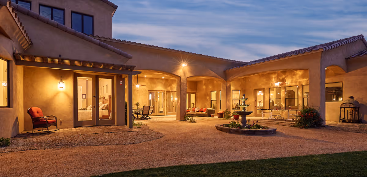 Evening view of an outdoor courtyard area at Abellavida Memory Care facility, featuring warm lighting, a central water fountain, seating areas with chairs and tables, and a building with stucco walls and tiled roof surrounding the courtyard.