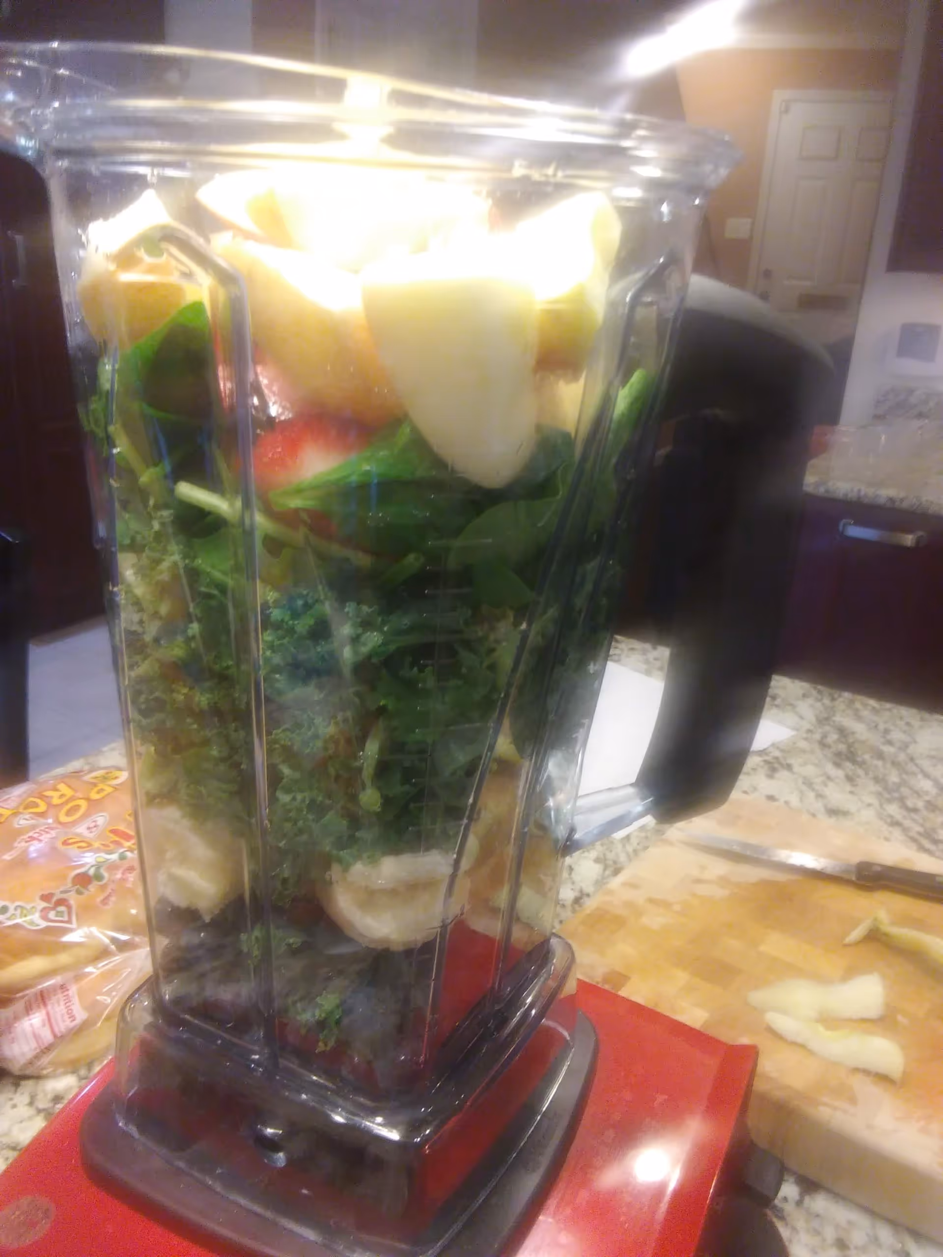 A blender filled with various fresh ingredients including leafy greens, banana slices, and other fruits, placed on a kitchen countertop with a cutting board and knife nearby.