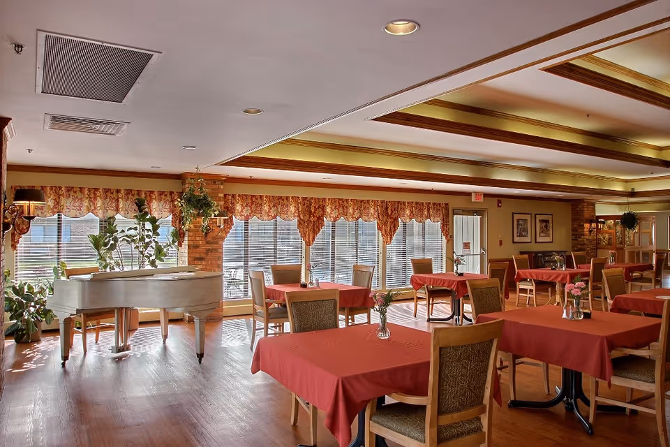 A spacious dining room with multiple tables covered in red tablecloths, each set with small flower vases. The room features large windows with floral valances allowing natural light to fill the space. There is a white grand piano on the left side near some green plants, and the ceiling has recessed lighting with wooden beams.