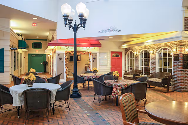 Indoor common area designed to look like an outdoor street scene with brick flooring, a black street lamp with three lights, several round tables covered with floral tablecloths and surrounded by wicker chairs, and a red door with windows and benches along the walls.