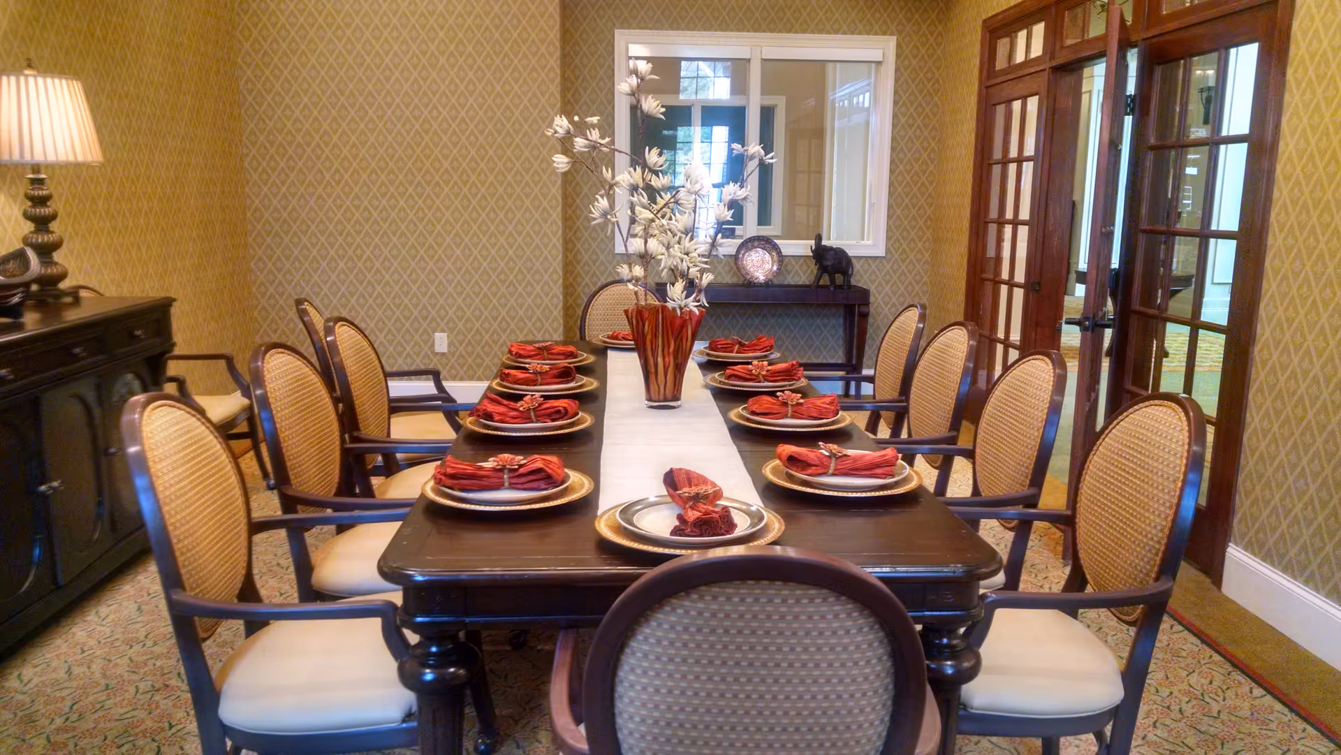 Formal dining room with a long table set with plates, red napkins, a centerpiece, and upholstered chairs.