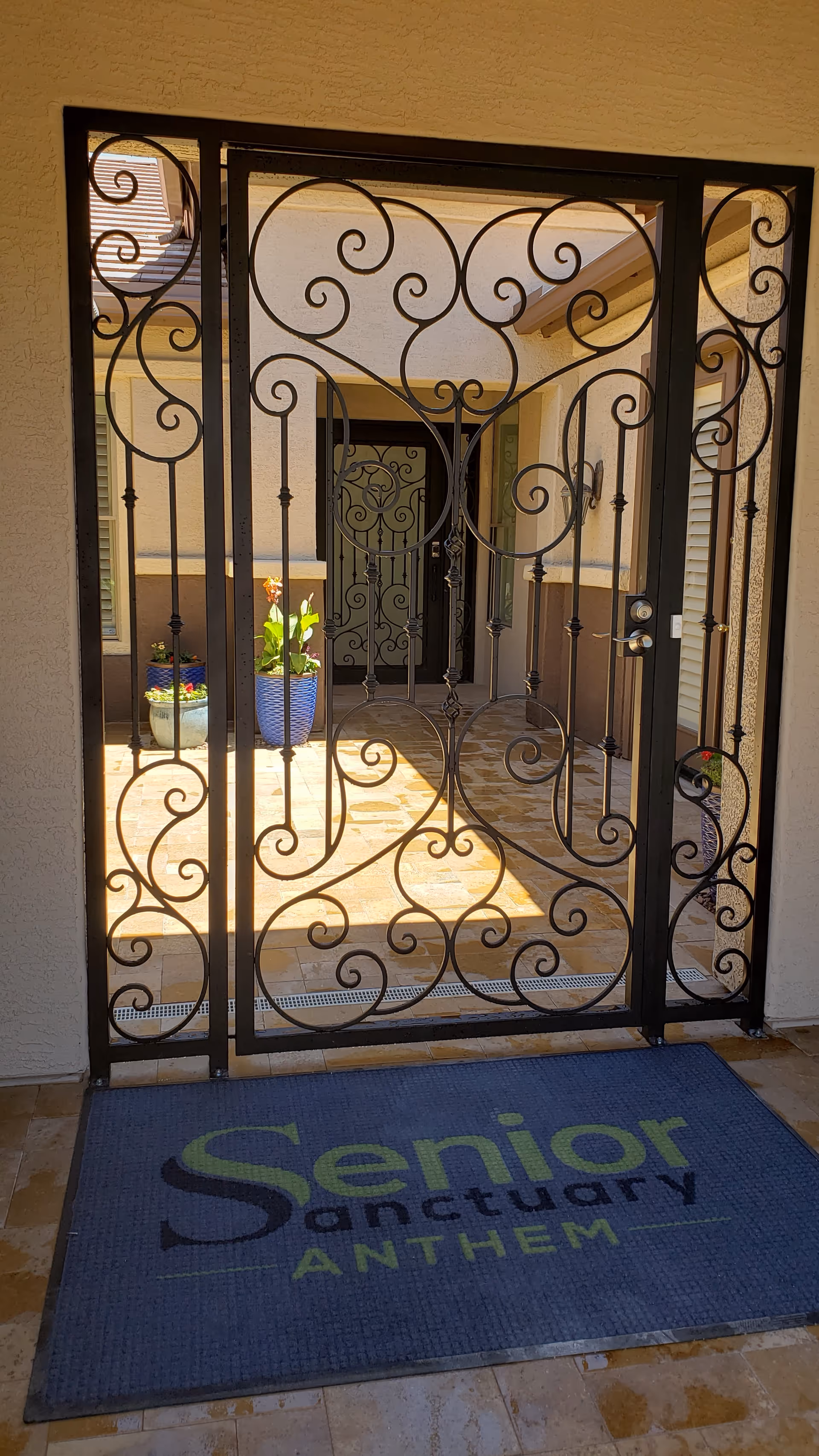 Ornate black metal gate with intricate scrollwork design at the entrance of a building. Behind the gate, there is a tiled courtyard with potted plants and a door with a similar decorative metal design. A blue doormat with the text 'Senior Sanctuary ANTHEM' is placed in front of the gate.