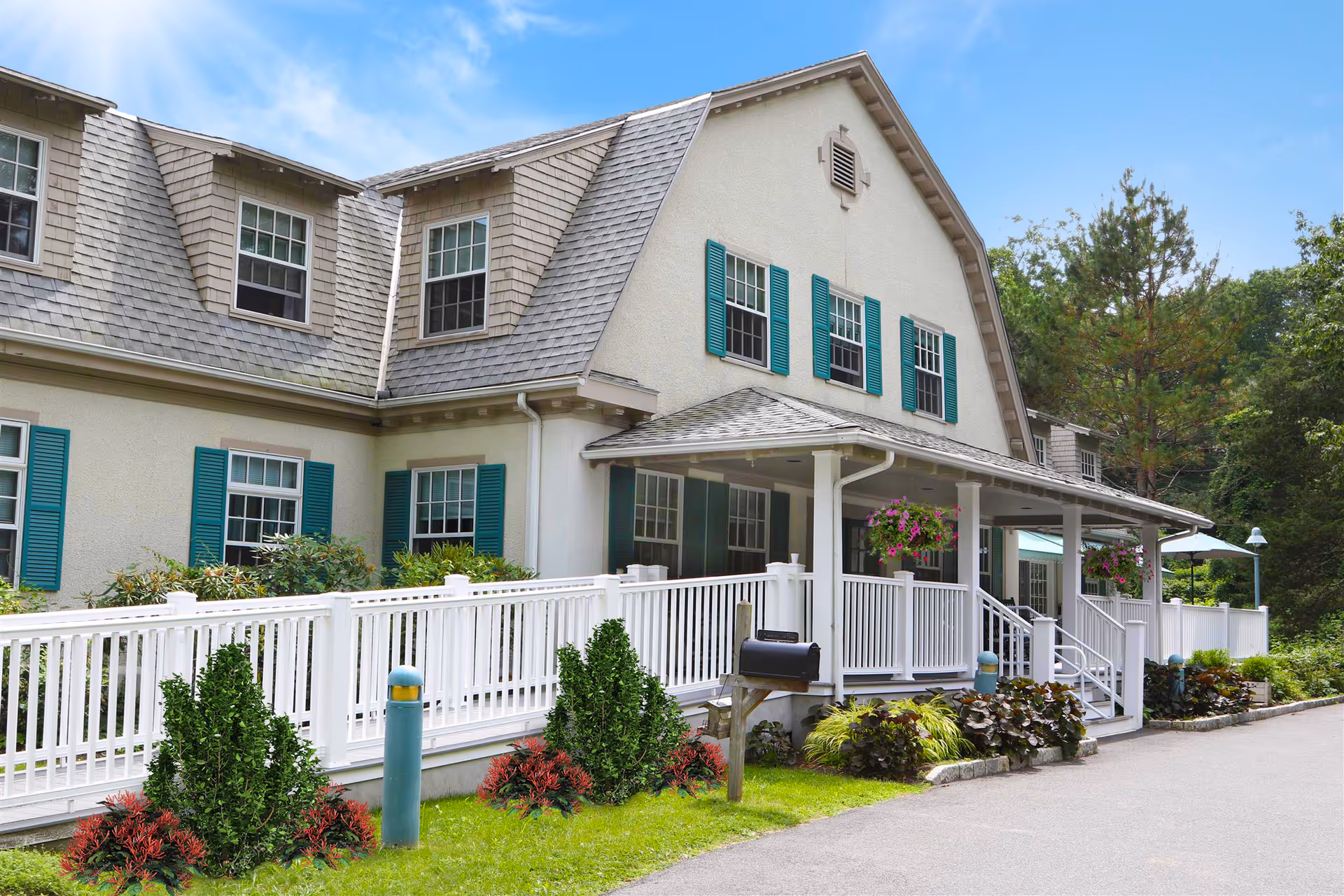 Exterior view of Parsonage Cottage Senior Residence showing a large, two-story building with beige walls, gray shingled roof, and teal window shutters. The building has a white railing along a ramp leading to a covered porch with hanging flower baskets. There is a mailbox on a wooden post near the entrance, surrounded by green shrubs and plants. The sky is clear and blue with some light clouds.