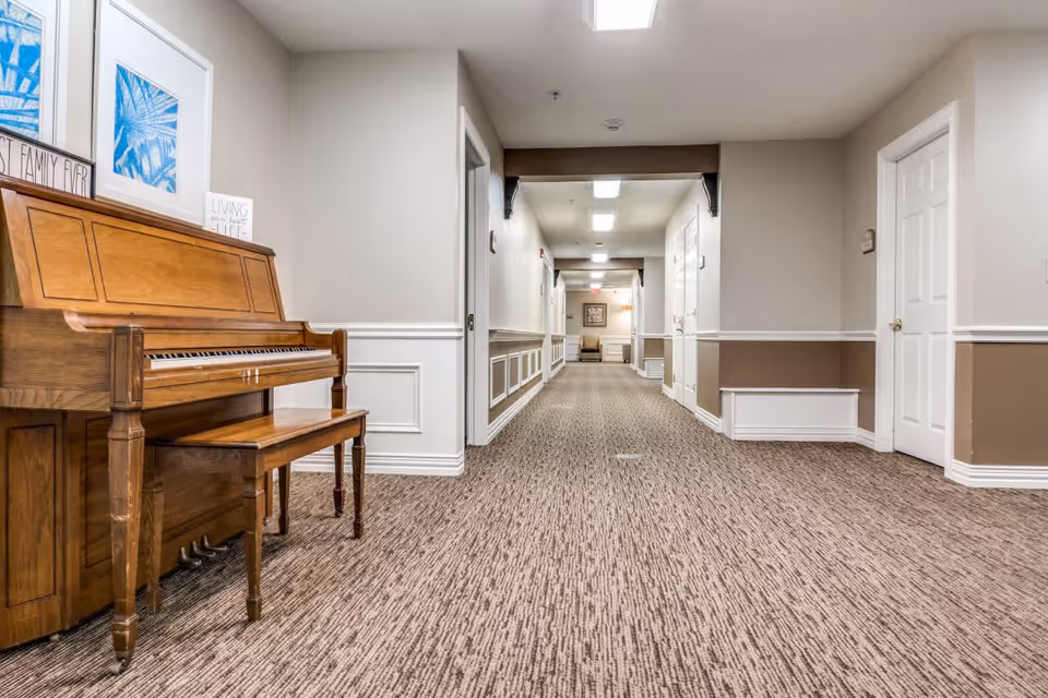 Carpeted interior corridor of a senior living facility with a wooden piano and bench on the left.