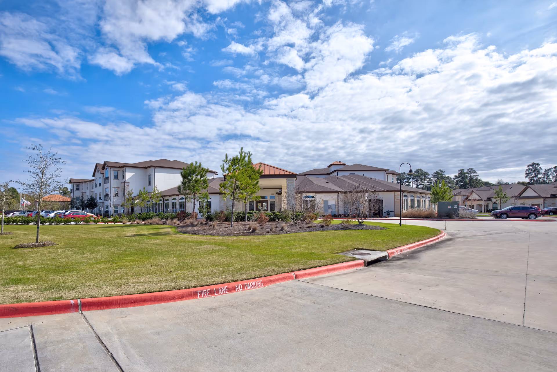 Front exterior of a senior living facility with a circular driveway, landscaped lawn, and a partly cloudy sky.