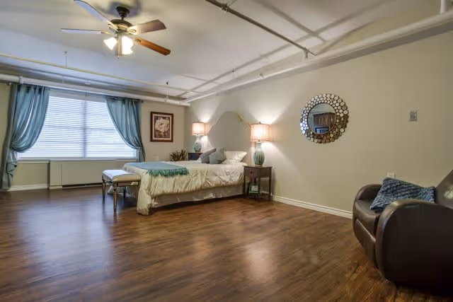 A spacious bedroom with a single bed against the wall, two bedside tables with lamps, a decorative round mirror on the wall, a large window with blue curtains, a ceiling fan with lights, a framed picture on the wall, a bench at the foot of the bed, and a dark armchair with a blue cushion on the right side.
