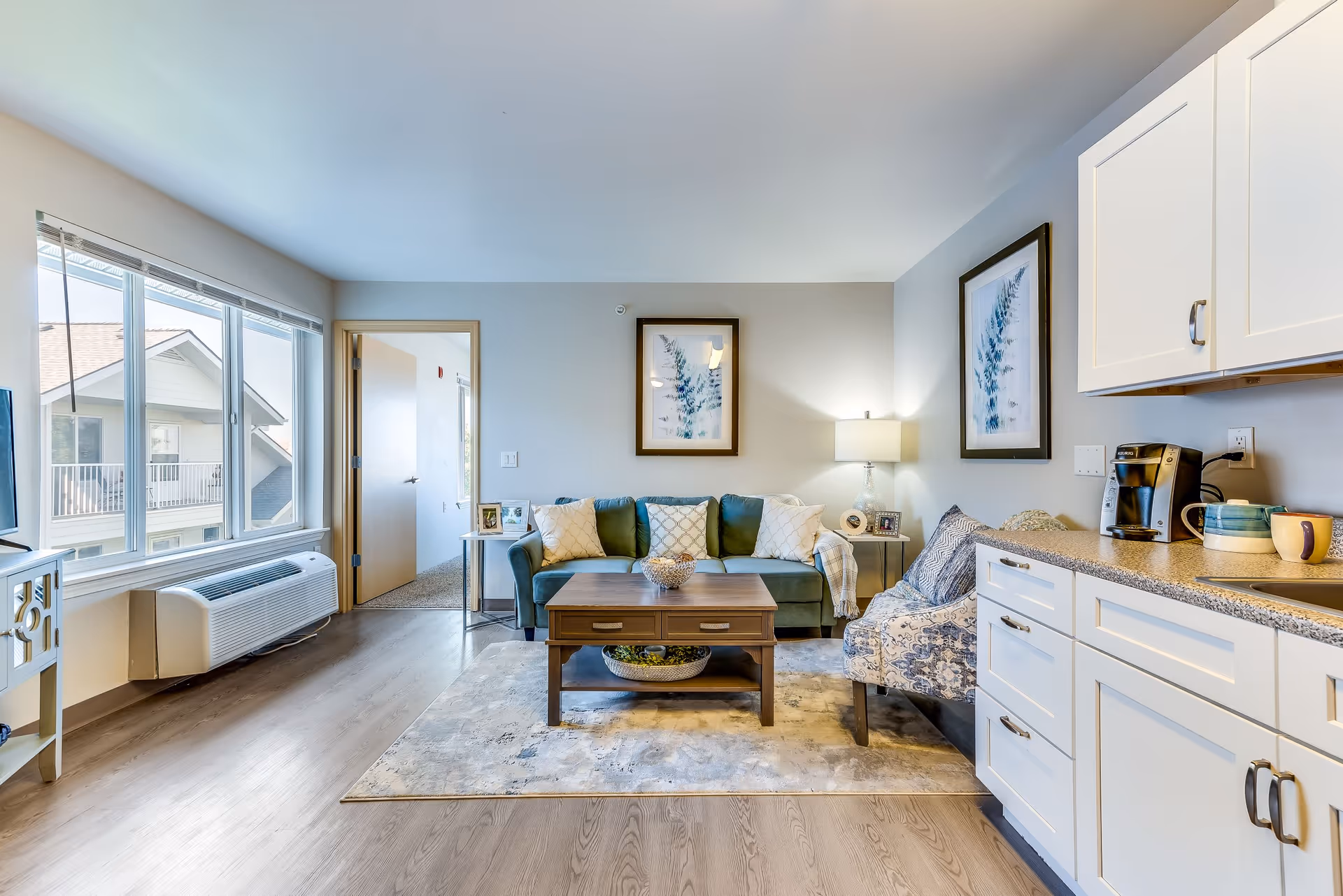 A bright and cozy living room area in a senior living facility with a green sofa adorned with decorative pillows, a wooden coffee table on a patterned rug, a patterned armchair, and white cabinetry with a coffee maker on the right. Large windows on the left let in natural light, and framed botanical artwork hangs on the walls.